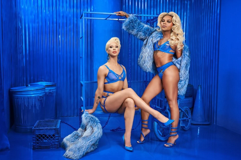 Two adult drag queens pose indoors in matching blue lingerie and fur coats against a matching blue set. The performers sit and stand.