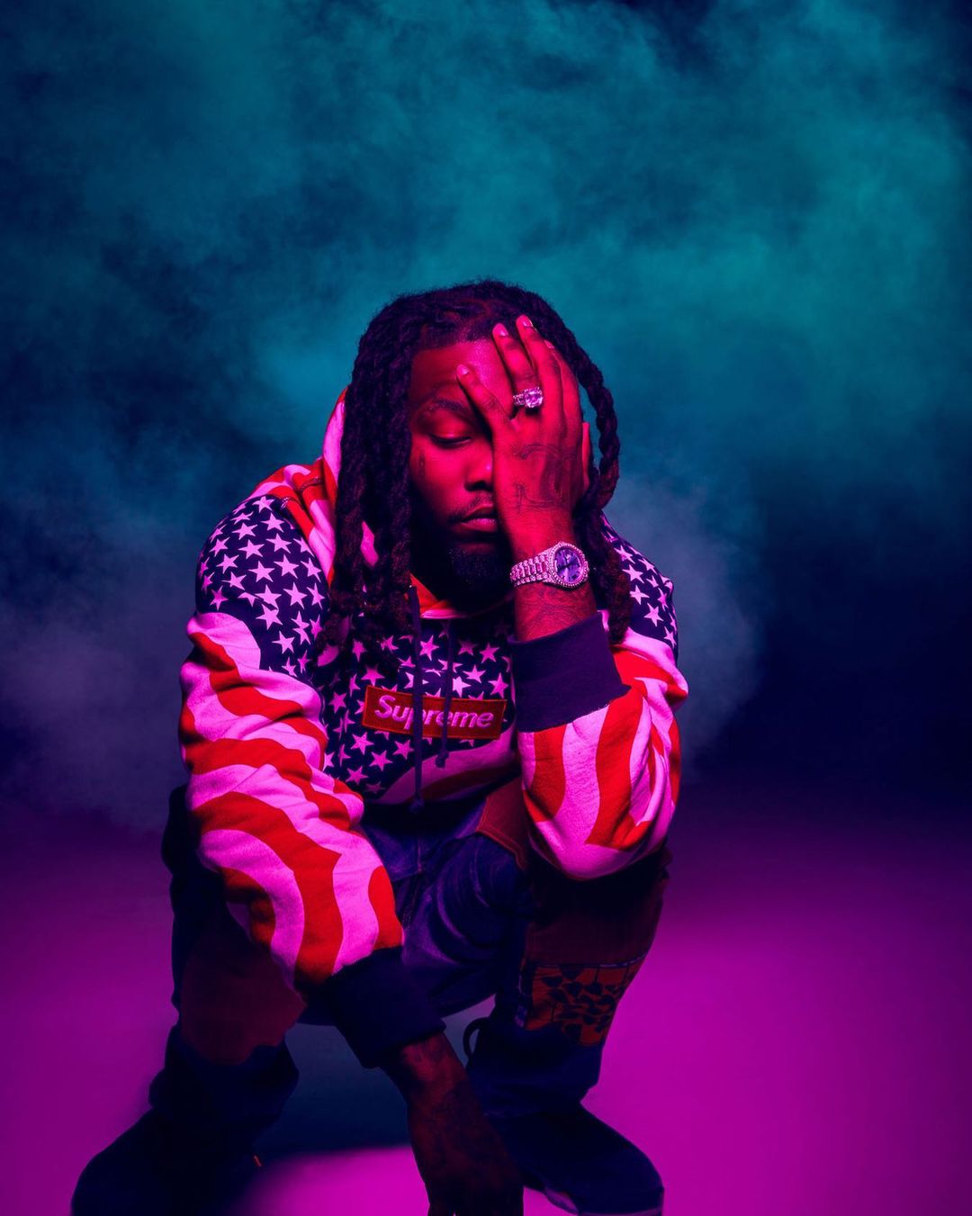 An adult male in a hoodie with an American flag pattern is crouching indoors with a hand covering part of his face, set against a smoky background.