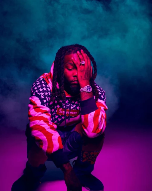 An adult male in a hoodie with an American flag pattern is crouching indoors with a hand covering part of his face, set against a smoky background.