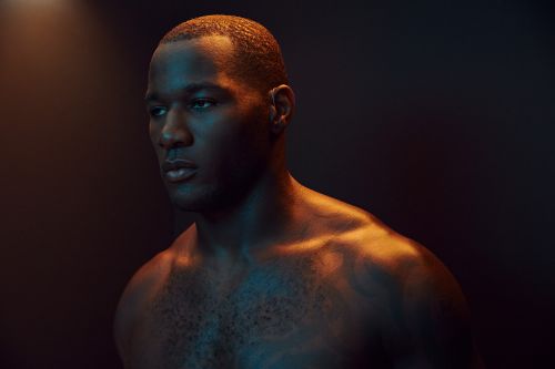 Striking portrait of a muscular Black man with a shaved head, showcasing athletic build and dramatic orange and blue lighting against a dark backdrop.