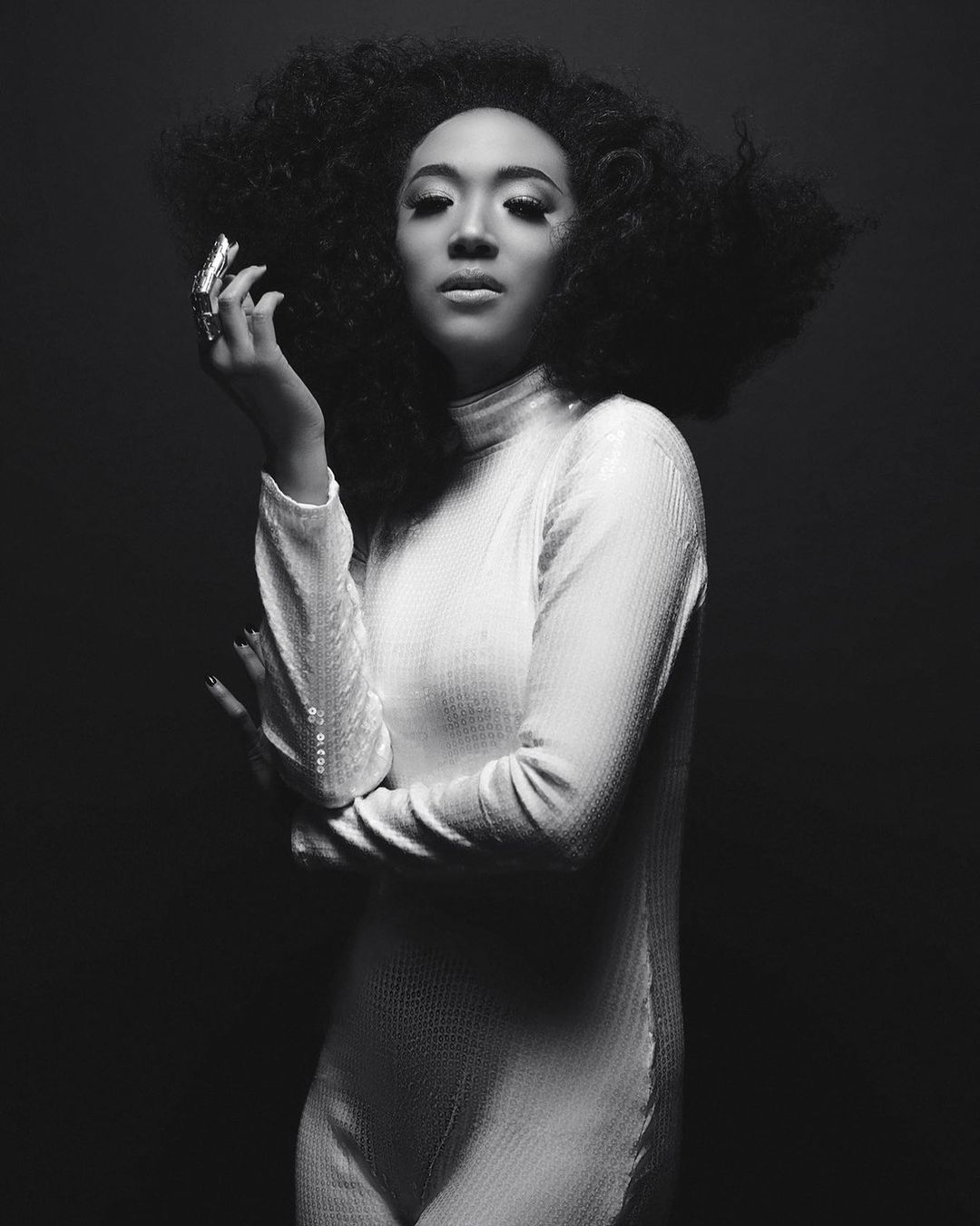 Monochrome portrait of a woman with an afro wearing a high-necked dress, looking directly at the camera in a studio setting.