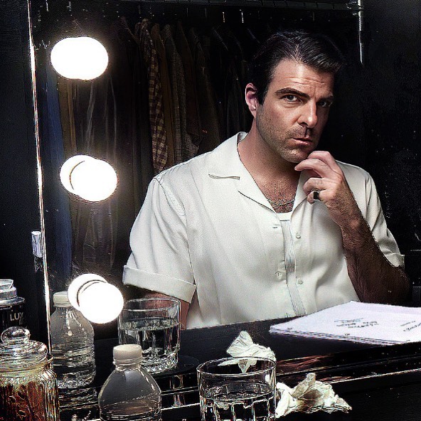 Portrait of an adult male with dark hair and a white shirt, seated at a table with a script and glasses of water in a dressing room.
