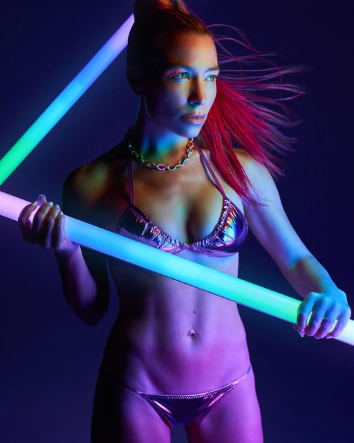 Woman with red-streaked hair in metallic bikini posing with neon lights against a dark background in a beauty-style studio shot.