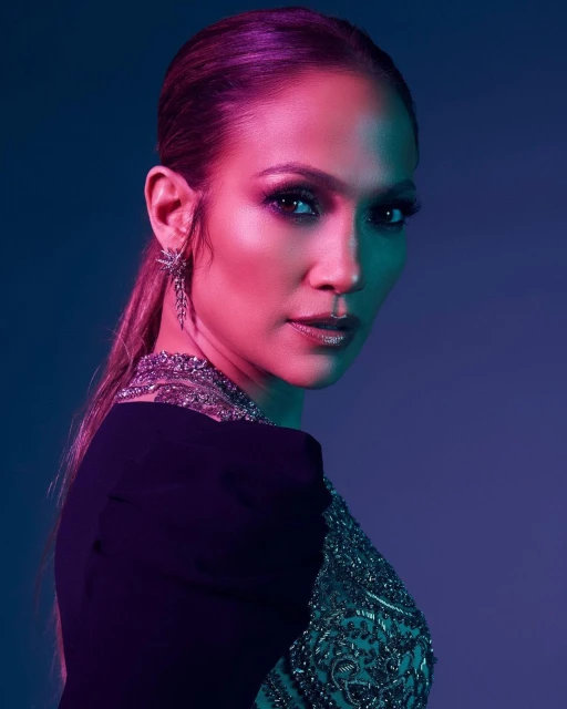 Close-up of adult female with long brown hair, wearing a black dress with silver beaded embellishments, posing against a blue backdrop.