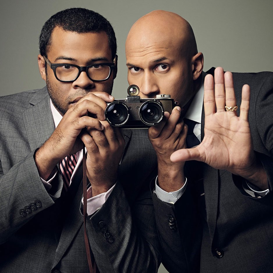 Two men in suits pose creatively with a vintage camera, one holding his hand up. Sharp studio portrait on a neutral background.