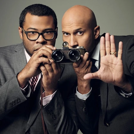 Two men in suits pose creatively with a vintage camera, one holding his hand up. Sharp studio portrait on a neutral background.