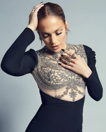 Jennifer Lopez poses in a black long-sleeved dress with intricate silver beading. She has her hands near her face and wears multiple rings.