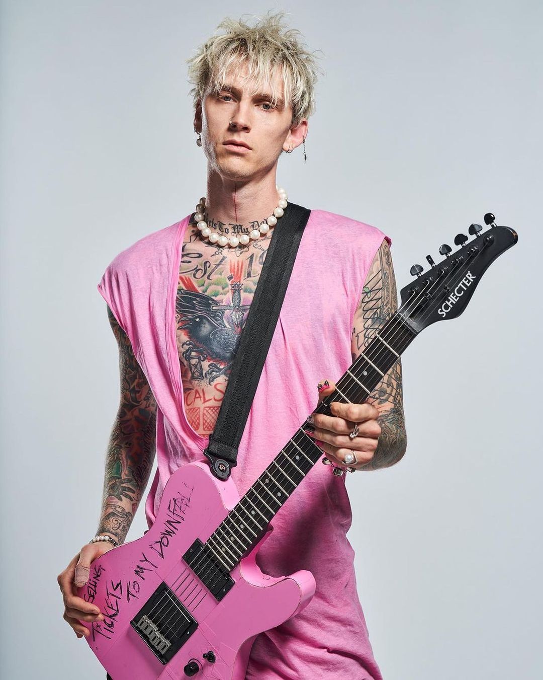 Machine Gun Kelly poses with a pink Schecter guitar, wearing a sleeveless pink shirt and pearl necklace, against a neutral studio backdrop.