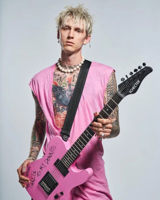Machine Gun Kelly poses with a pink Schecter guitar, wearing a sleeveless pink shirt and pearl necklace, against a neutral studio backdrop.