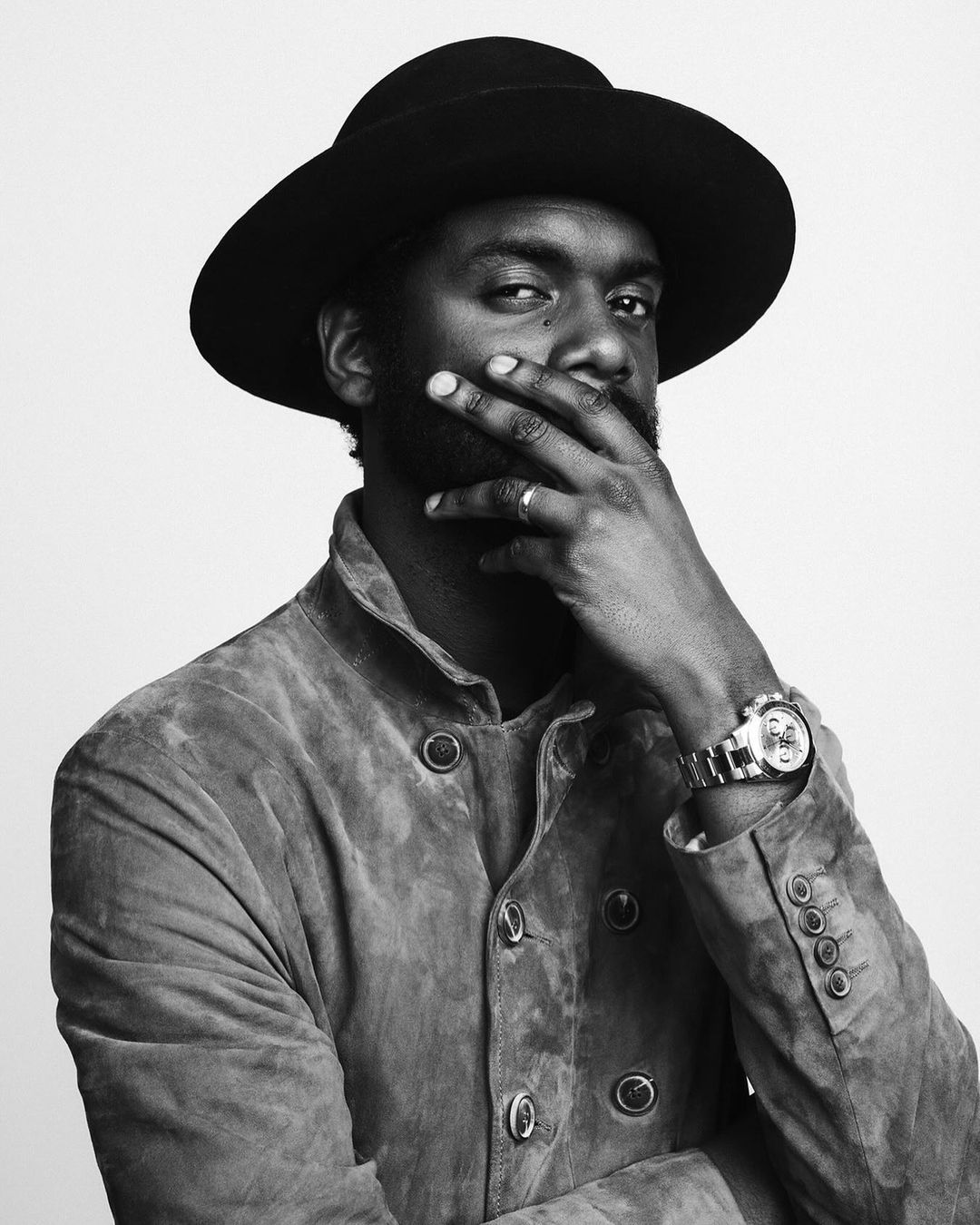 Striking black and white portrait of a bearded adult male in a hat and jacket, hand to face, watch visible, against a plain backdrop.