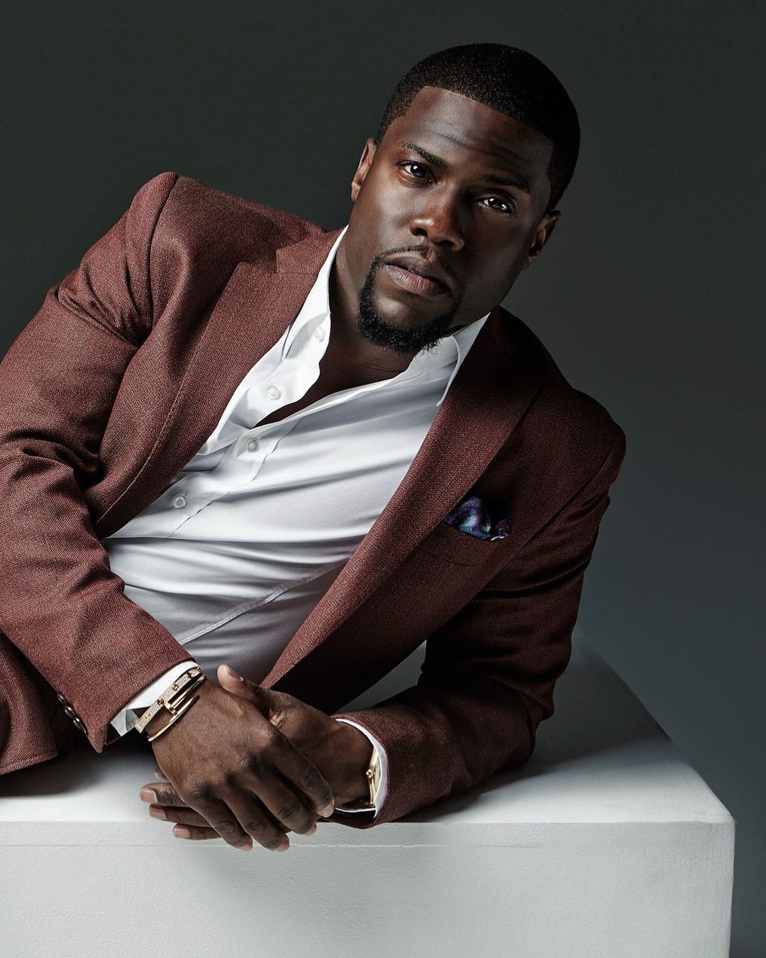Headshot of an adult male with dark skin, wearing a maroon suit with a white dress shirt, sitting against a gray gradient background.