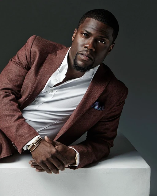 Headshot of an adult male with dark skin, wearing a maroon suit with a white dress shirt, sitting against a gray gradient background.