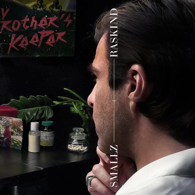 Side profile portrait of a thoughtful adult male next to bottles, plants, and a framed photograph, against a dark background.
