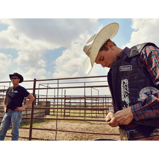 Two men in cowboy hats near a rodeo arena on a sunny day with clouds. One man wears a protective vest over a plaid shirt.