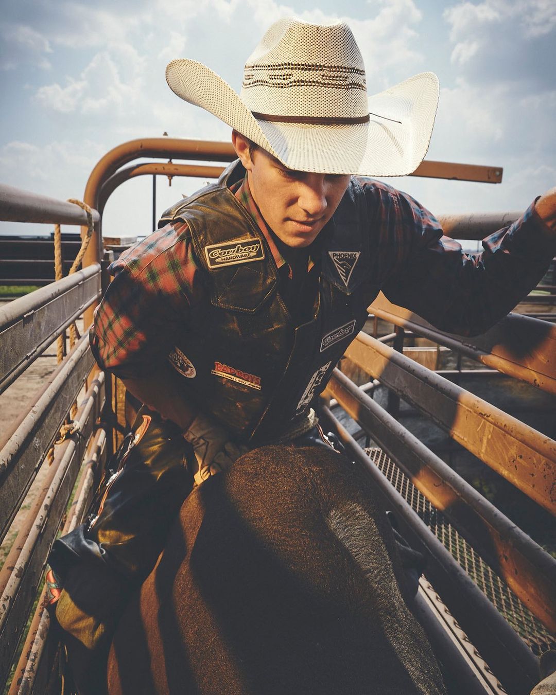 Cowboy on a bull inside a rodeo pen, wearing a straw hat and a protective vest, with a metal gate and clouded sky background.