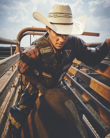 Cowboy on a bull inside a rodeo pen, wearing a straw hat and a protective vest, with a metal gate and clouded sky background.