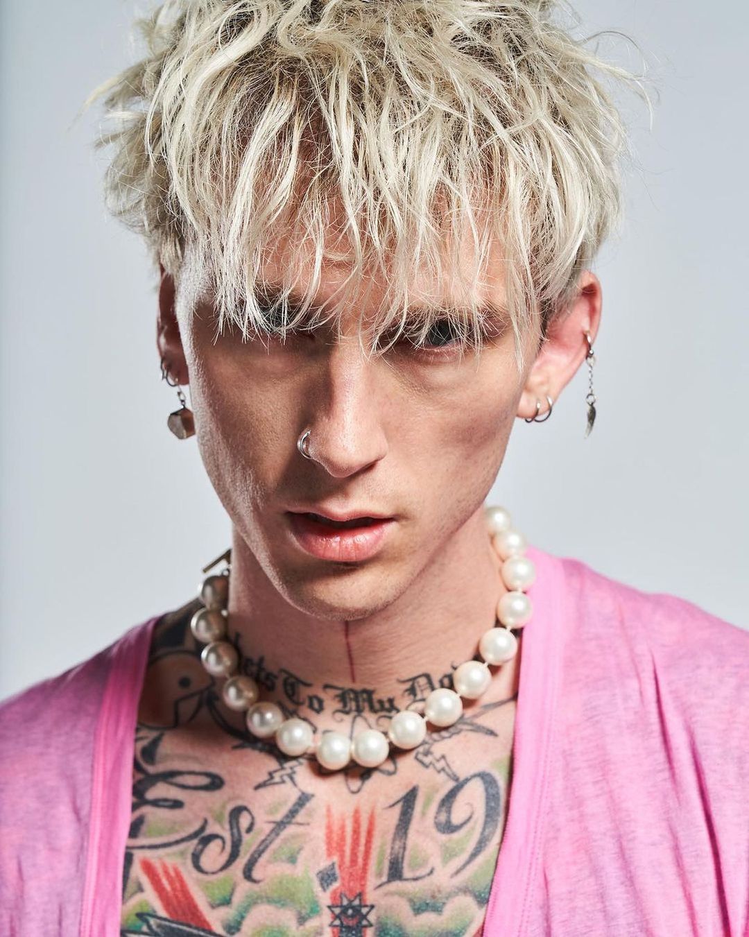 Close-up portrait of a blond adult male in a pink shirt and pearl necklace, showcasing tattoos against a neutral background.