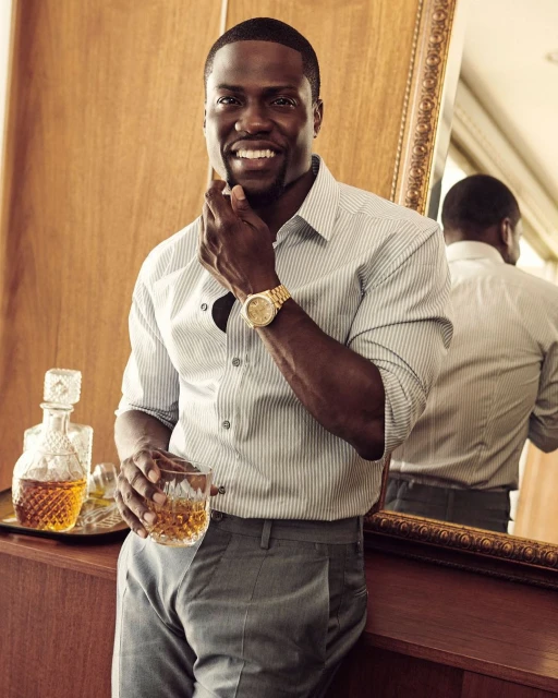 Portrait of smiling adult Black male in grey pants and striped shirt, gold watch, holding a glass of whiskey indoors.