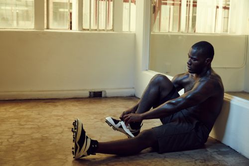 Dark-skinned male athlete sitting on a floor, tying the laces of his football shoes in an indoor training space with natural light.
