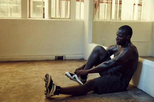 Dark-skinned male athlete sitting on a floor, tying the laces of his football shoes in an indoor training space with natural light.