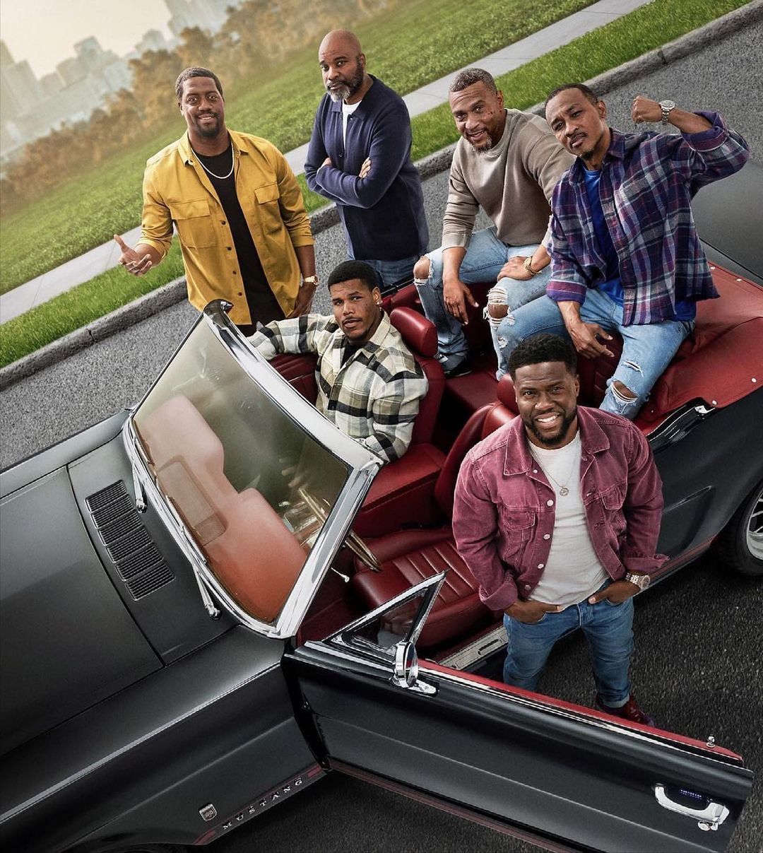 Group of six African American men pose with a classic black convertible car on a paved street with green trees in the background.