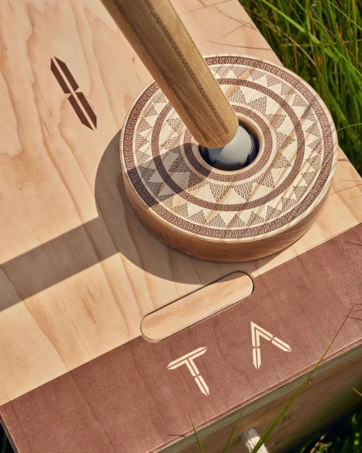 Overhead shot of a wooden game board with decorative concentric circles and a wooden rod in a grassy outdoor setting.