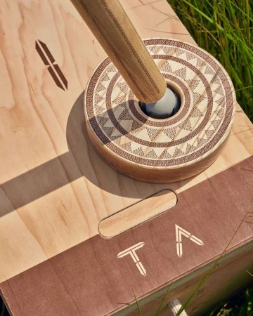 Overhead shot of a wooden game board with decorative concentric circles and a wooden rod in a grassy outdoor setting.