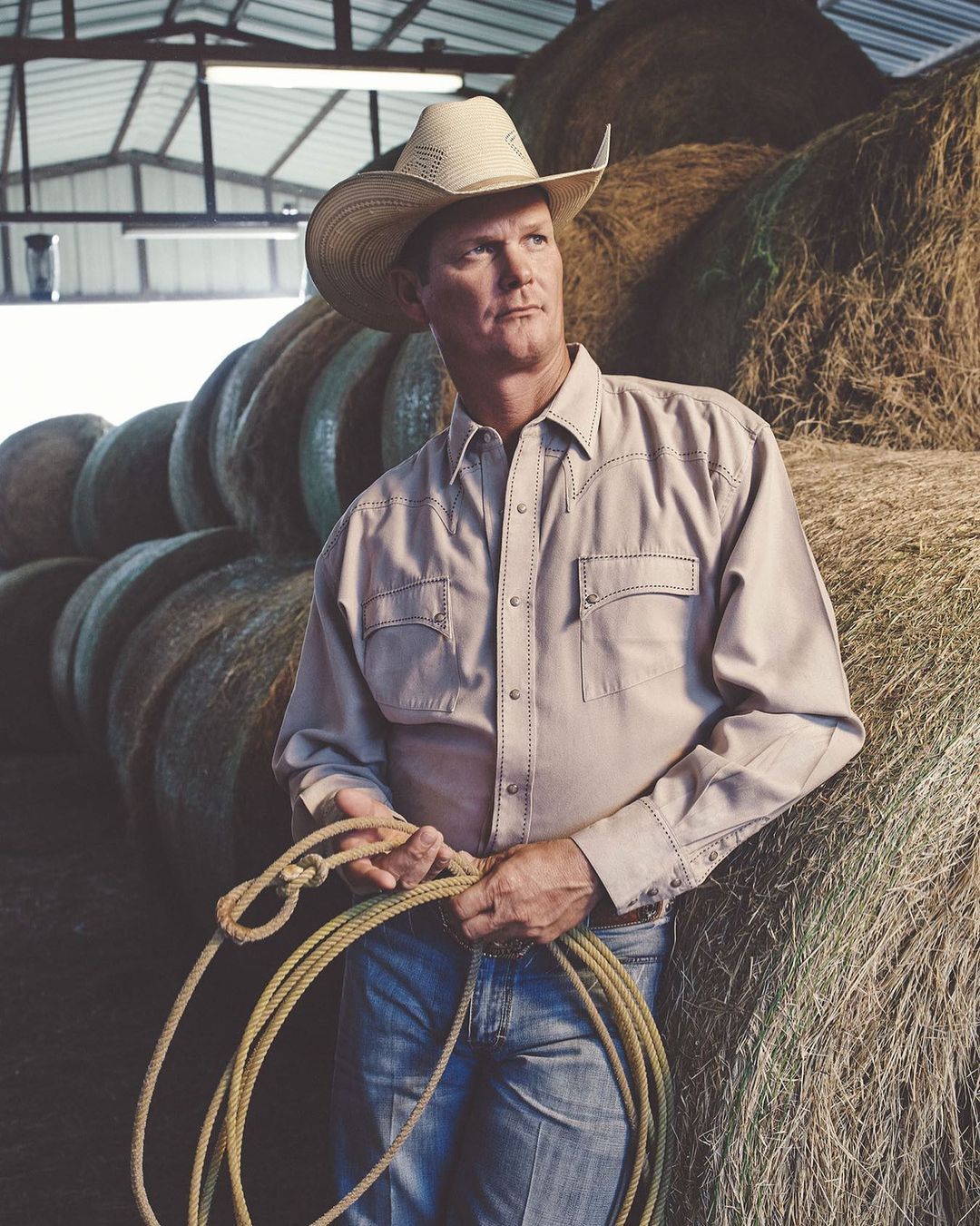 Man in cowboy hat poses with rope in barn with hay bales, wearing a beige shirt and jeans, looking off into the distance.