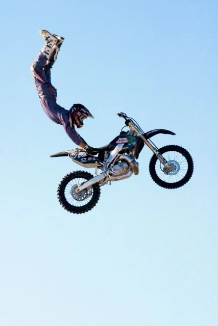 Motocross rider performs a stunt in mid-air on a dirt bike against a clear sky; the rider is upside down while holding onto the bike.