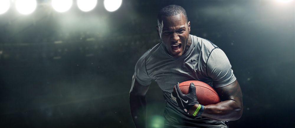 Intense action shot of an African-American football player gripping the ball tightly, face contorted in a yell, bathed in stadium lights.