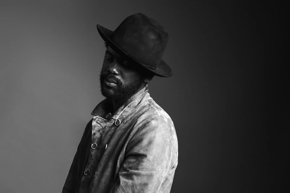 Black and white studio portrait of an adult male wearing a fedora and jacket against a gradient gray background.
