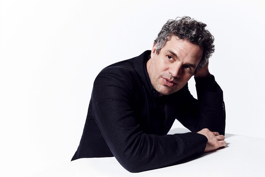 Portrait of a mature adult male with graying curly hair, wearing a dark suit jacket, posing in a photo studio setting against a white backdrop.