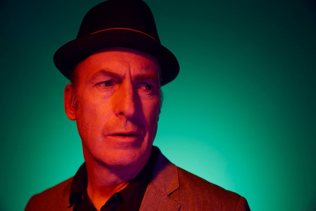Close-up portrait of a serious-looking man wearing a dark hat and a grey suit jacket against a vibrant green background.