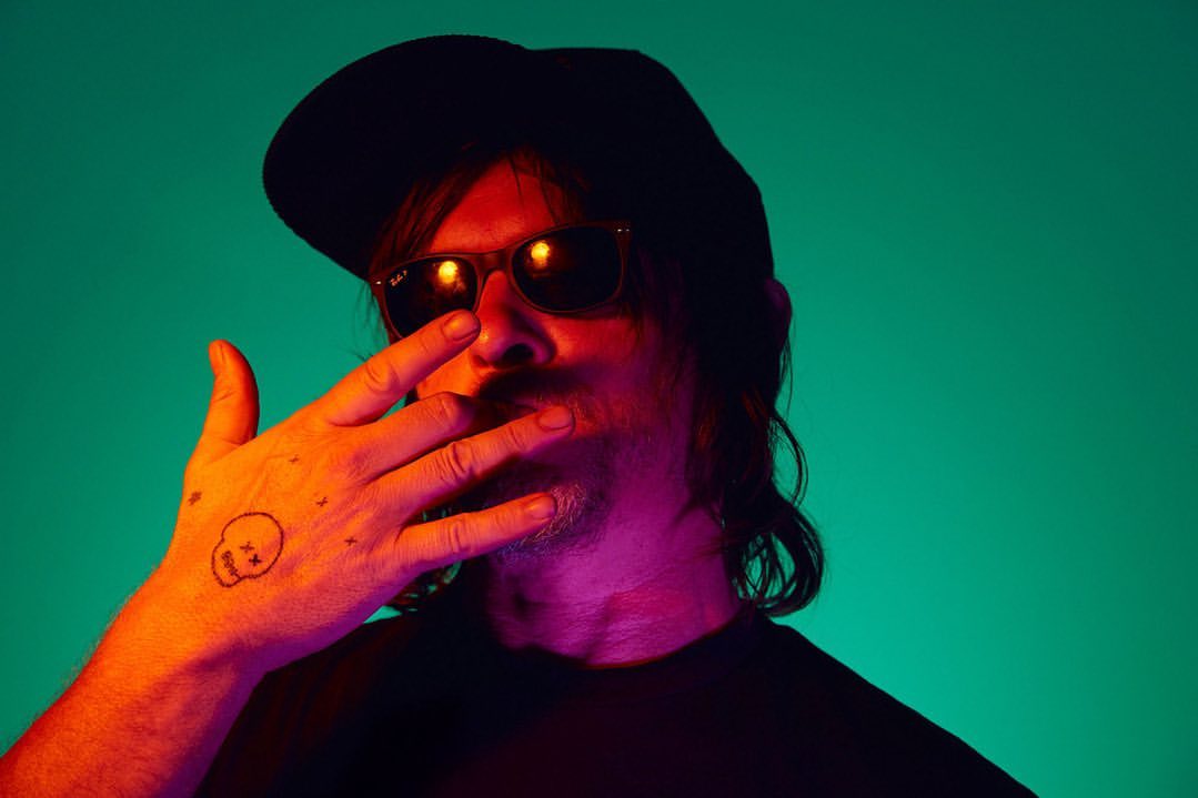 Portrait of adult male in sunglasses and black cap, set against vibrant teal and orange lighting, with a hand tattoo detail.