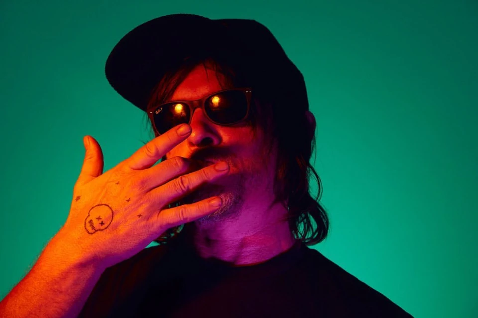 Portrait of adult male in sunglasses and black cap, set against vibrant teal and orange lighting, with a hand tattoo detail.