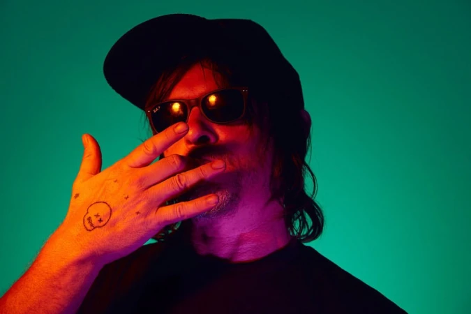 Portrait of adult male in sunglasses and black cap, set against vibrant teal and orange lighting, with a hand tattoo detail.