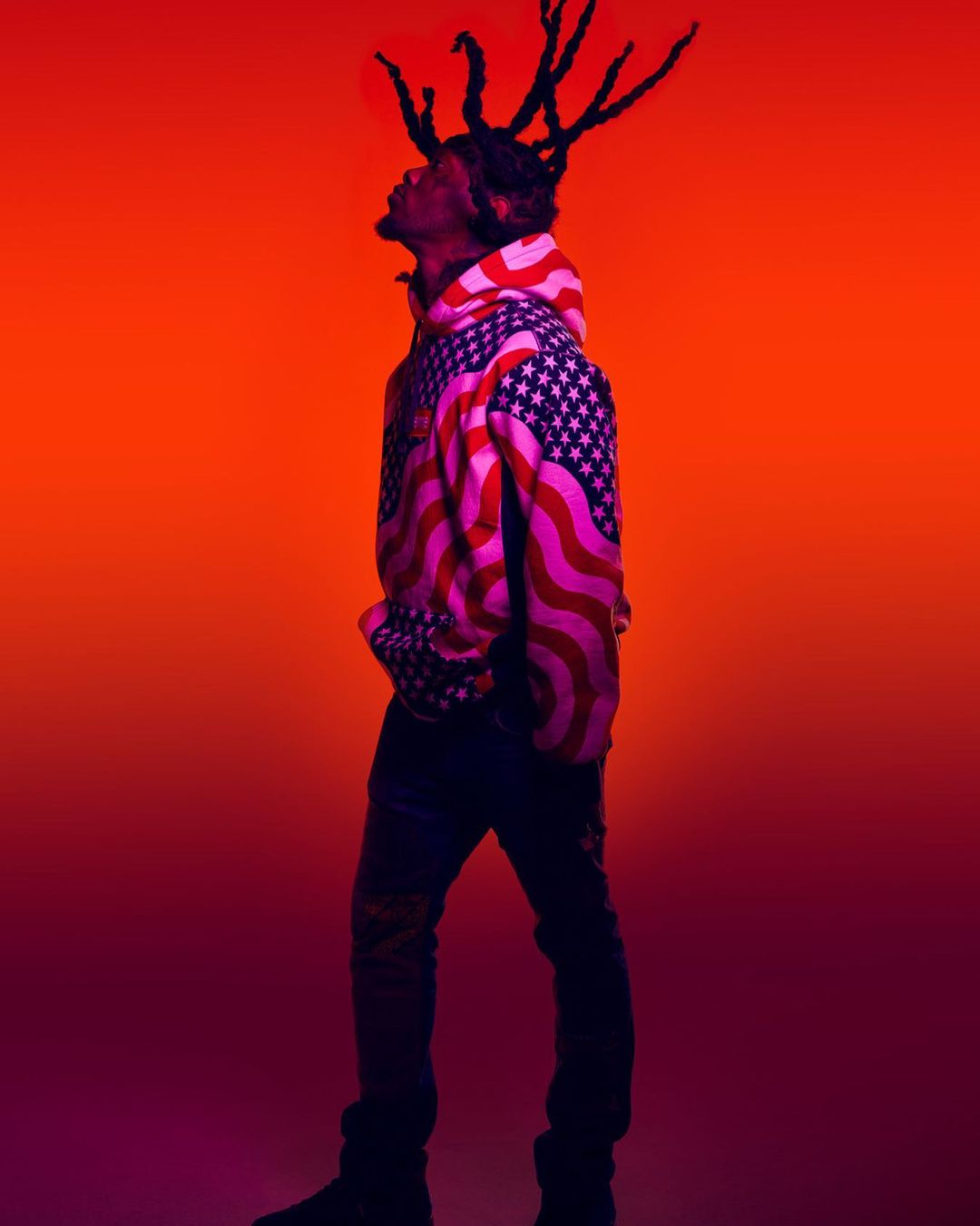 Adult male in hooded sweatshirt patterned with American flag stands against a vibrant red-orange gradient backdrop.