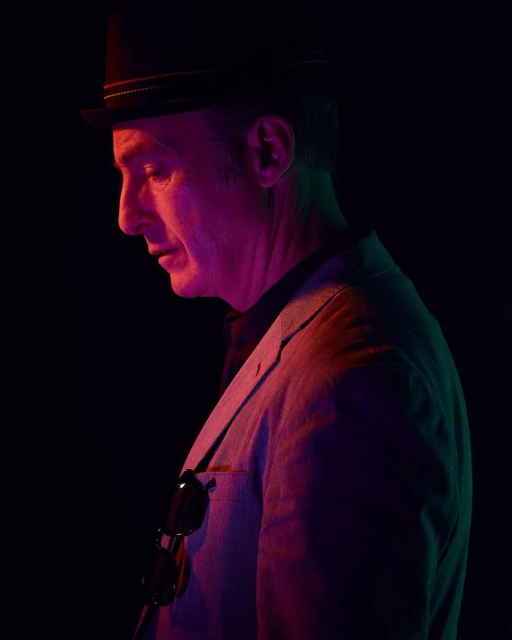 Profile portrait of a man in a fedora and jacket, lit with moody pink and green hues against a stark black background.