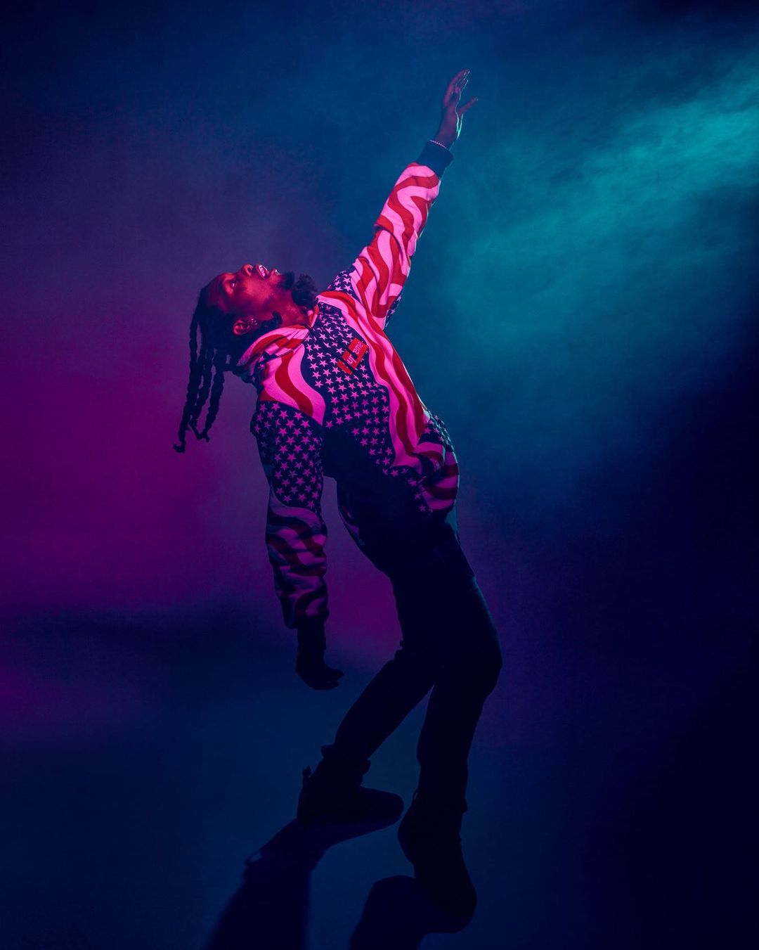 Man in American flag hoodie dancing under blue and purple stage lights with raised arm and dark pants and sneakers in studio.