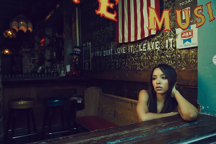 Indoor portrait of a young woman seated at a wooden booth near a bar, with an American flag and music sign in the background.