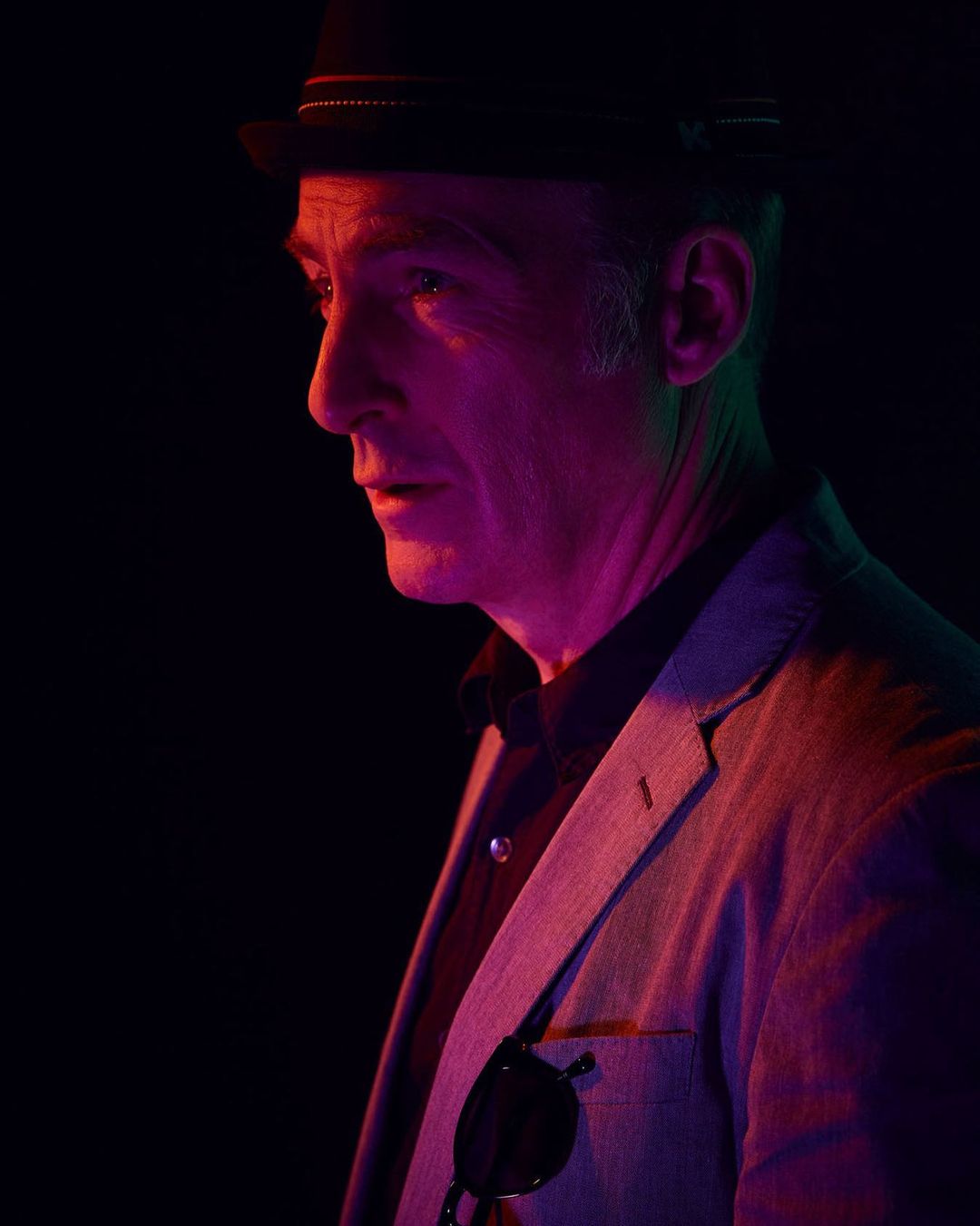 Portrait of a mature adult man in a hat and blazer, lit with pink and purple gel lighting against a dark background in a studio setting.