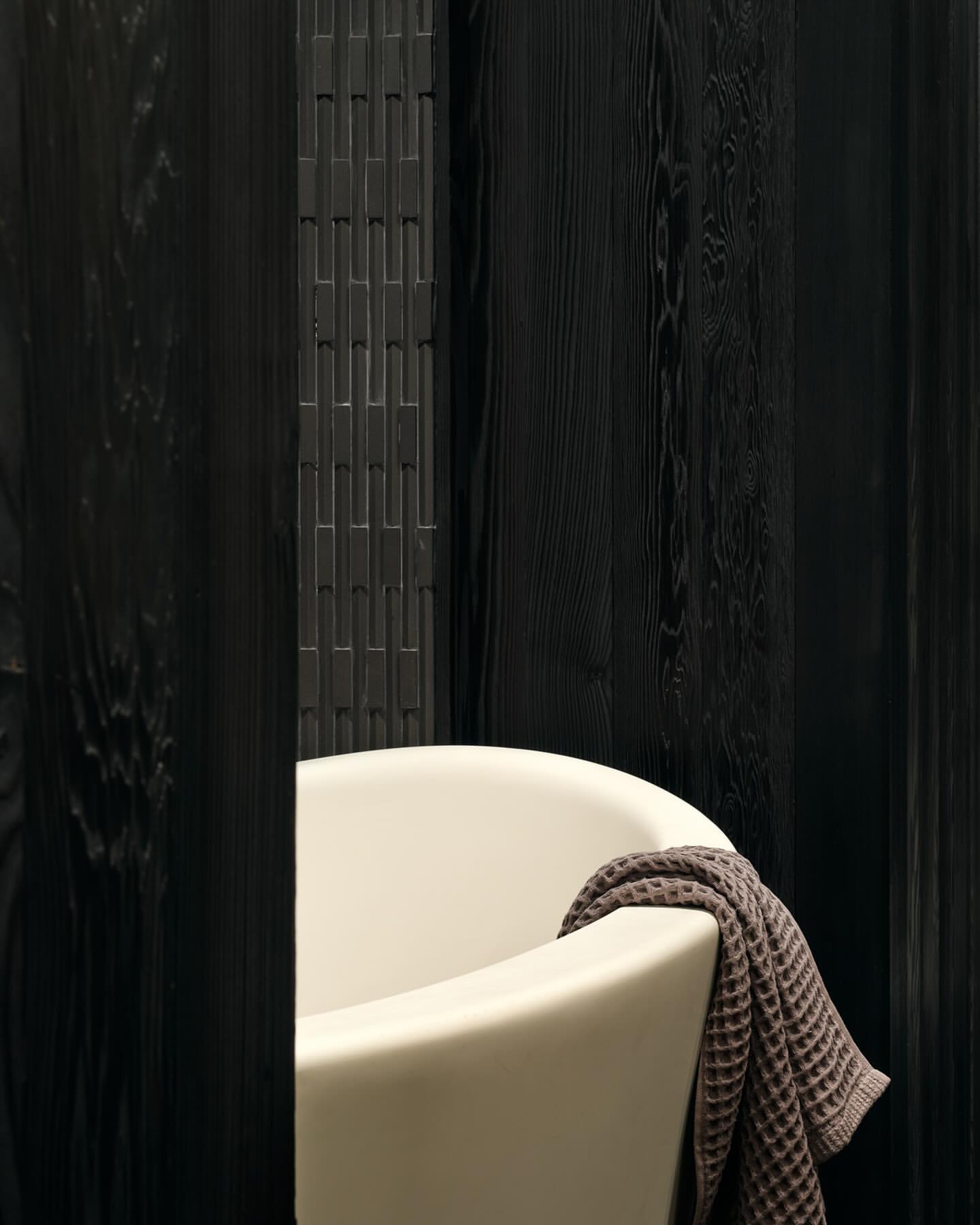 A cream-colored bathtub with a brown towel draped over the side, set against a backdrop of dark, vertical wood panels.