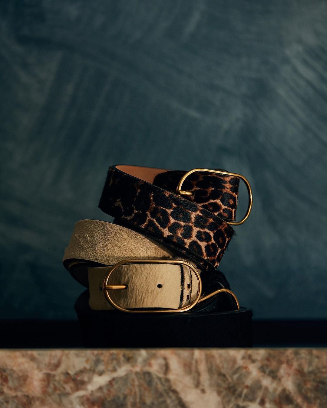 Stack of fashion belts: one animal print, one beige with gold buckles, on a marble surface against a textured dark blue background.