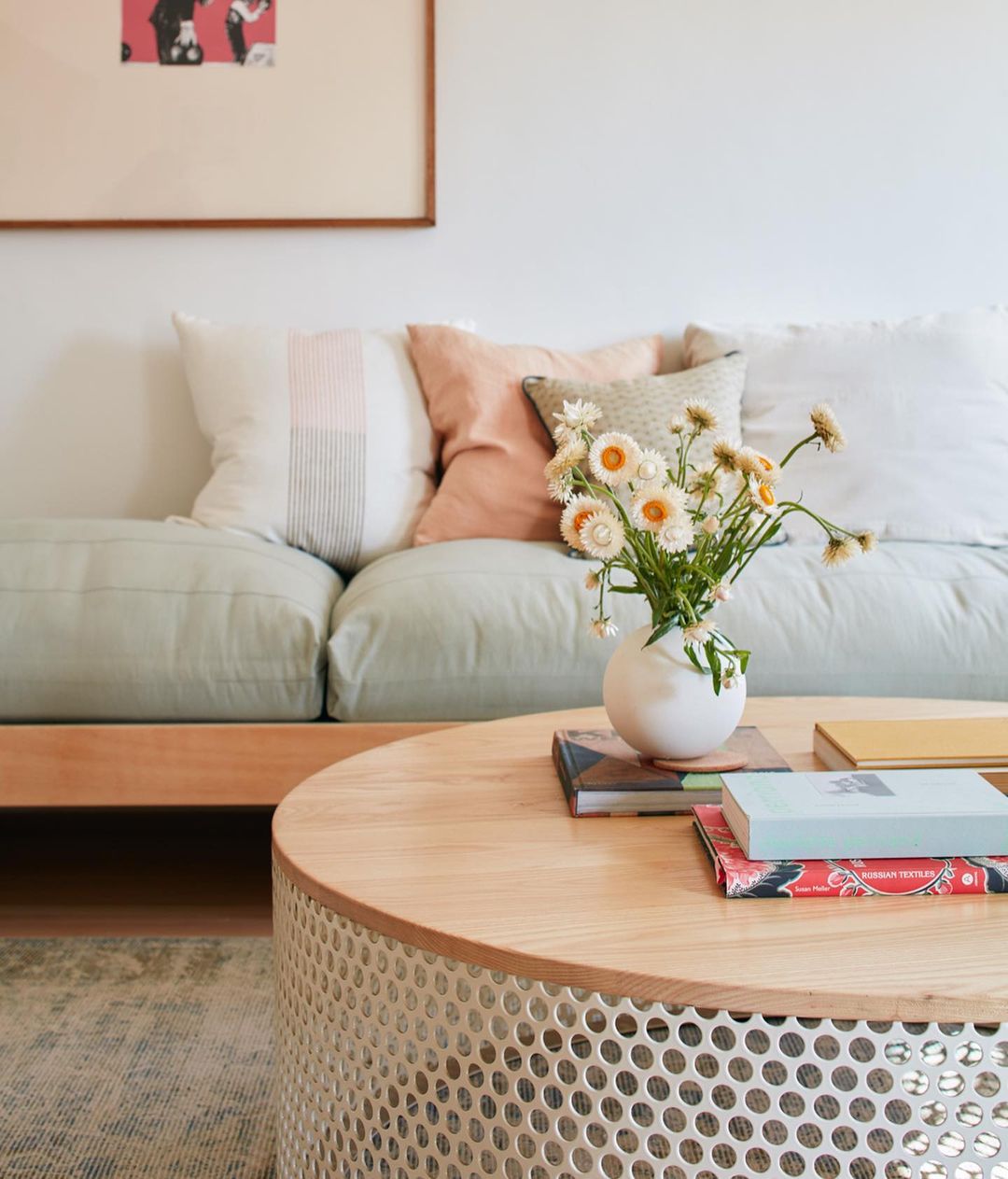 Living room interior: a round wood coffee table with a white vase of strawflowers, books, sits in front of a light green sofa with pillows.
