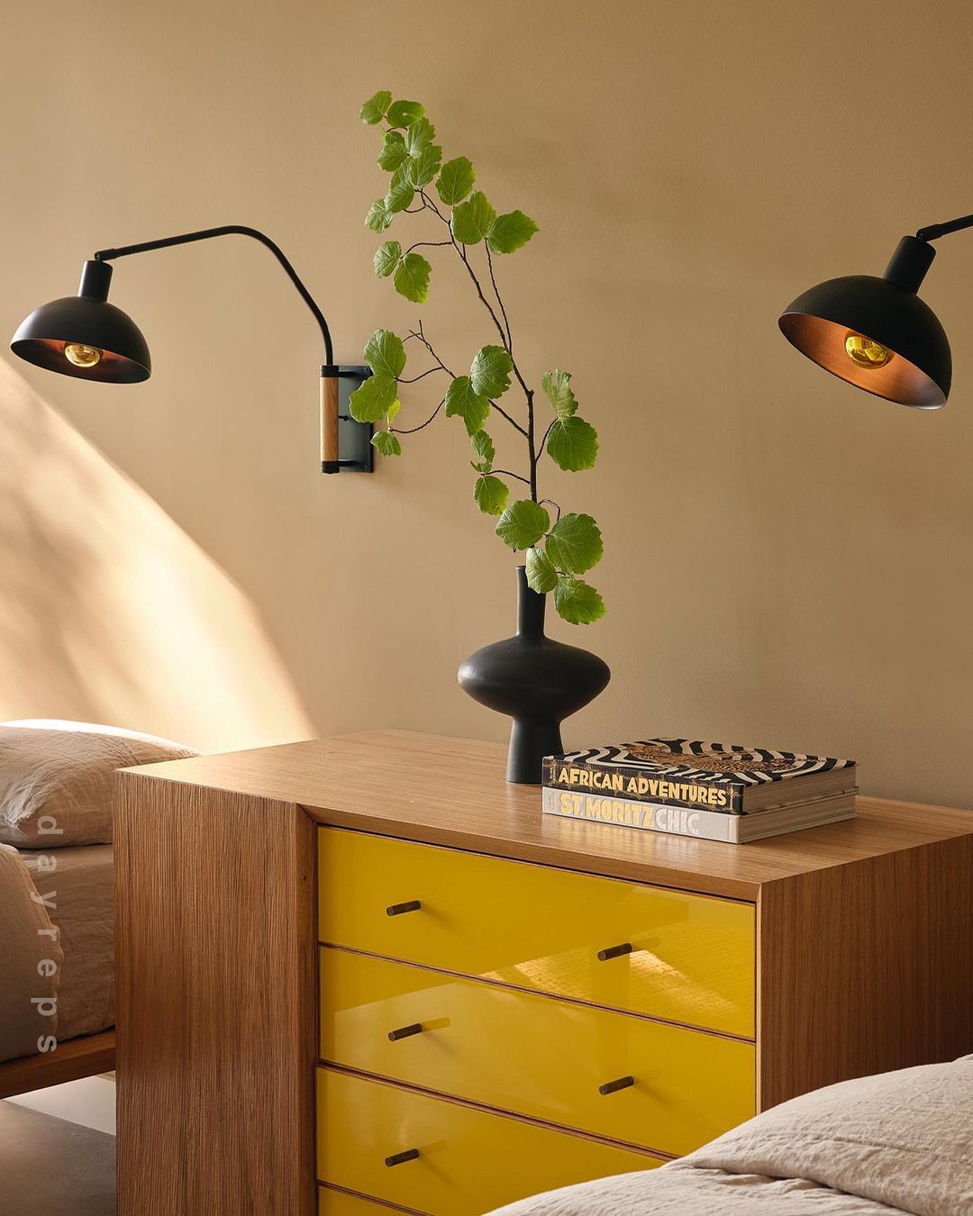 Bedroom interior featuring a yellow dresser with lamps, a black vase with greenery, and a book, creating a modern and stylish atmosphere.