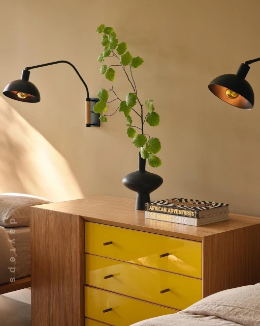 Bedroom interior featuring a yellow dresser with lamps, a black vase with greenery, and a book, creating a modern and stylish atmosphere.