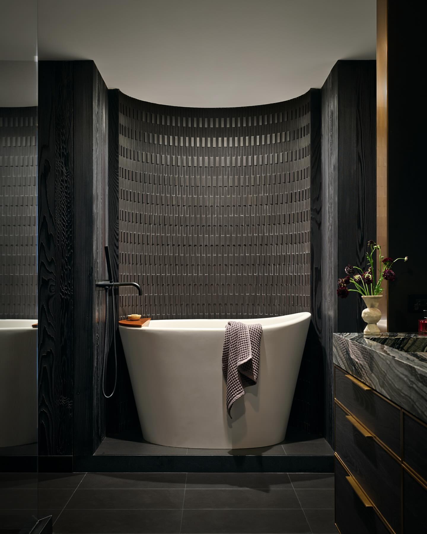 Modern bathroom features a white soaking tub with black wood wall panels and gray tile wall with black tile flooring indoors.