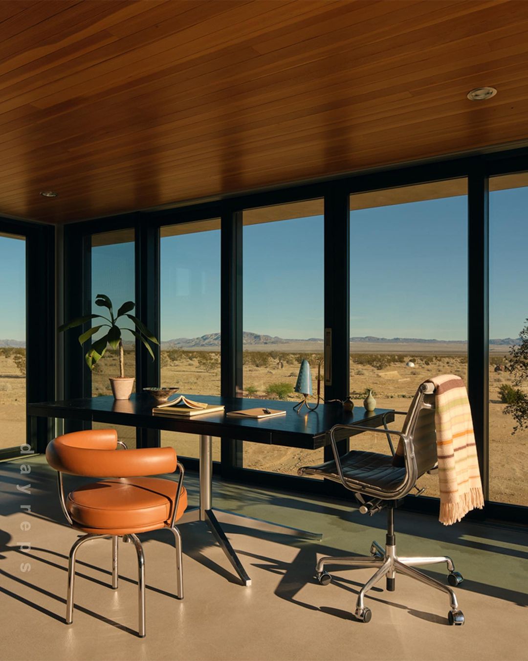 Home office with modern black desk and neutral-colored chairs in a room with large windows overlooking a desert landscape.