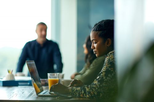 Woman working on a laptop at a table, with colleagues and a man standing in the background, creating a modern, collaborative workspace.