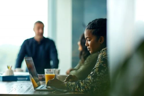 Woman working on a laptop at a table, with colleagues and a man standing in the background, creating a modern, collaborative workspace.